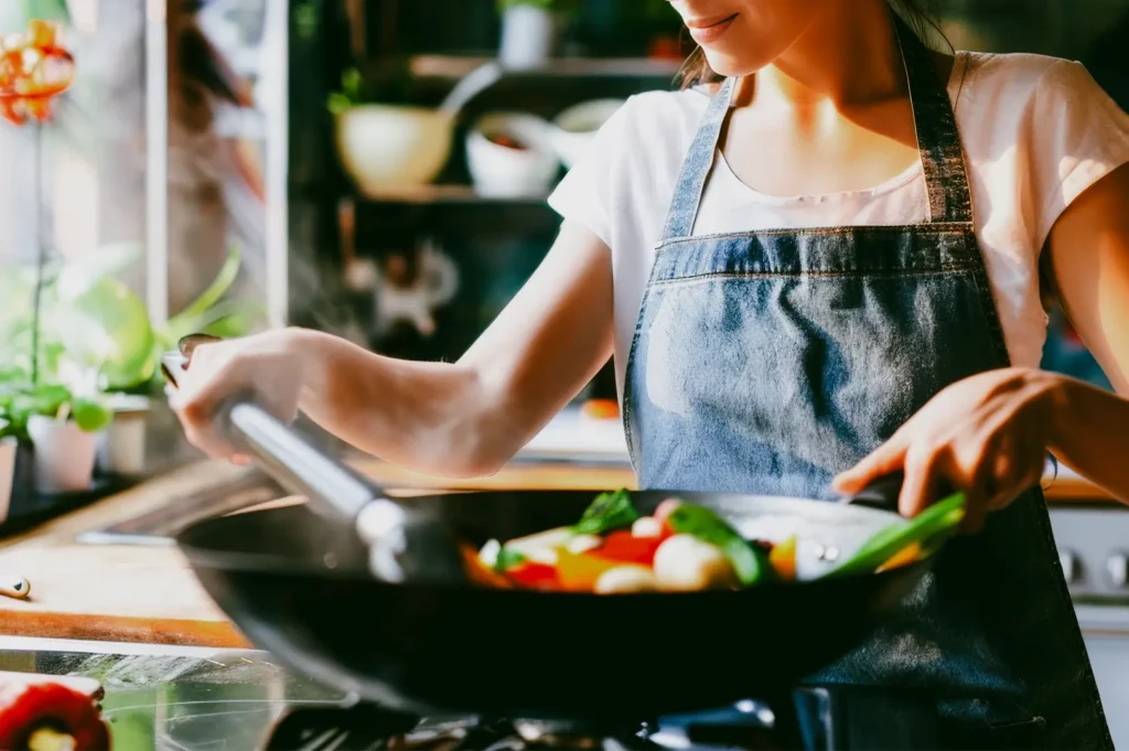 Side‑angle medium close‑up shot of a person stir‑frying fresh vegetables in a wok on a home kitchen stovetop, wearing an apron and cooking near a window with natural light.