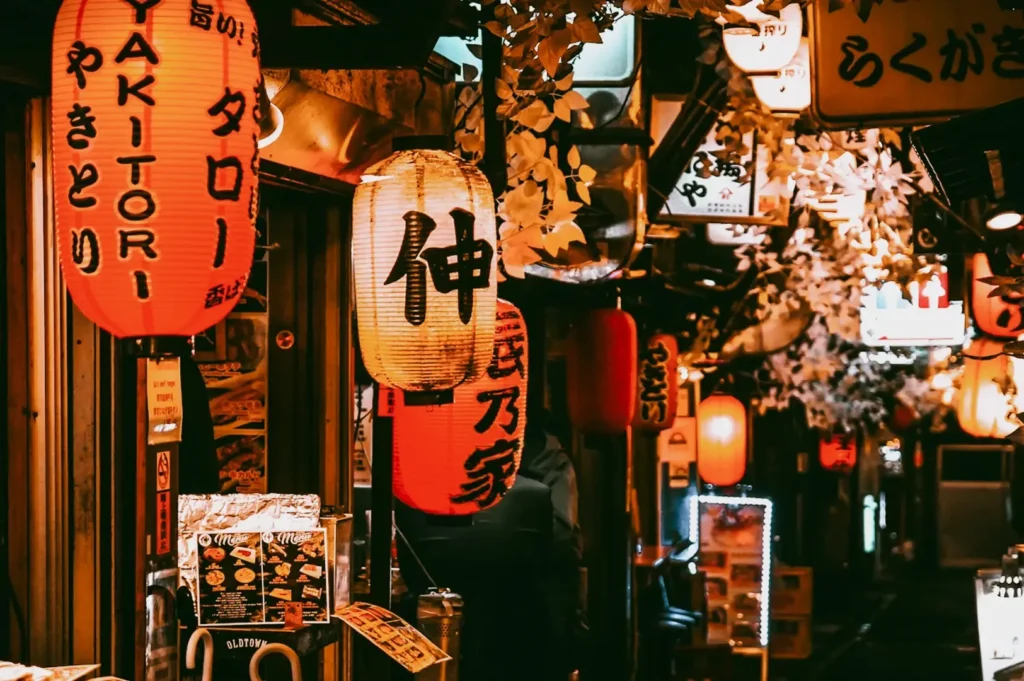 Eye-level, narrow-angle shot of a lantern-lined izakaya alley in Singapore, featuring multiple red, white, and warm amber paper lanterns printed with Japanese characters and words such as ‘yakitori,’ mounted closely along dark wooden storefronts with small menu boards and signage near the entrances; warm, low lighting casts an orange glow across textured walls, hanging foliage overhead, and illuminated sign boxes receding into the background, creating depth and a moody late-night izakaya atmosphere.