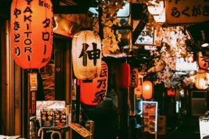 Eye-level, narrow-angle shot of a lantern-lined izakaya alley in Singapore, featuring multiple red, white, and warm amber paper lanterns printed with Japanese characters and words such as ‘yakitori,’ mounted closely along dark wooden storefronts with small menu boards and signage near the entrances; warm, low lighting casts an orange glow across textured walls, hanging foliage overhead, and illuminated sign boxes receding into the background, creating depth and a moody late-night izakaya atmosphere.