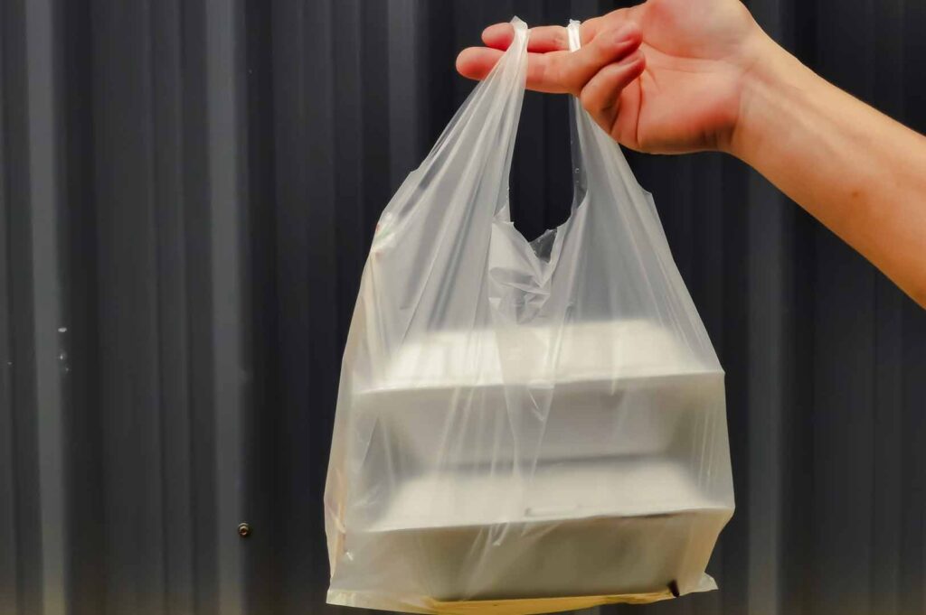 Hand holding a plastic bag containing stacked takeaway food containers against a dark background.