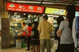 Wide-angle eye-level shot of Hua Kee Chicken Rice hawker stall in Singapore, featuring a red illuminated signboard with Chinese and English text, an open kitchen preparing Hainanese chicken rice, and a queue of customers waiting inside a traditional hawker centre setting.