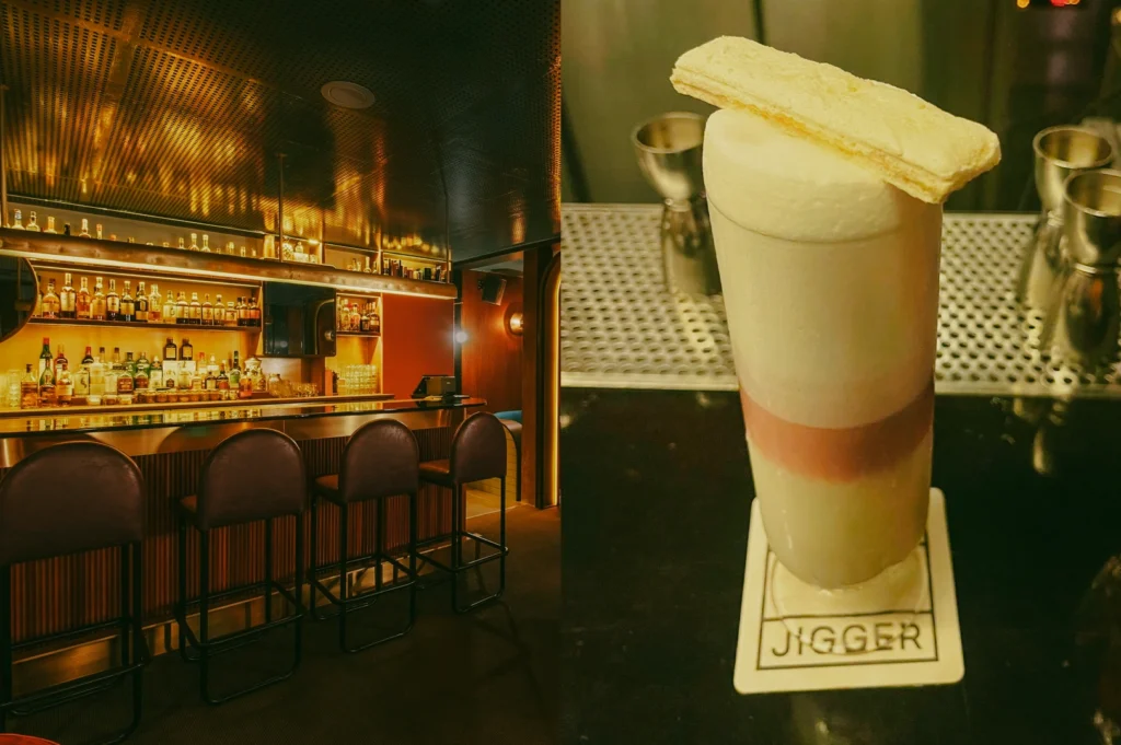 Left: A wide-angle, eye-level shot of a modern, upscale bar featuring a glossy countertop lined with four dark leather barstools. The back bar is heavily stocked with spirit bottles across illuminated shelves under a metallic, textured ceiling with warm, ambient lighting. Right: A close-up, eye-level shot of a tall cocktail glass layered with a light base, a pink middle band, and a thick white foam top, garnished with a long wafer biscuit. The glass rests on a square coaster bearing the text 'JIGGER' over a black reflective bar surface, with metal bar jiggers visible blurred in the background.