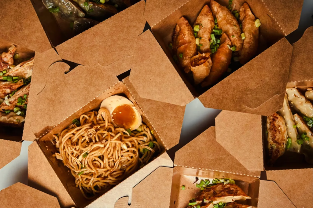 Top‑down view of multiple cardboard takeaway boxes filled with Asian dishes such as noodles, dumplings, and fried appetizers.
