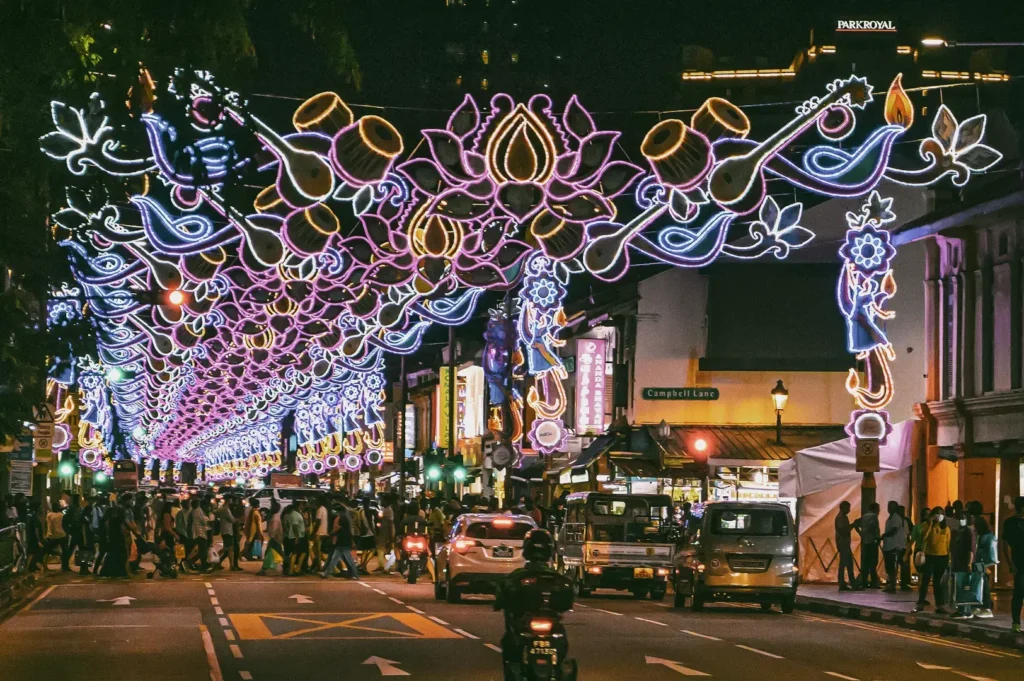 Wide-angle night shot of Little India in Singapore during Deepavali, with colorful light arches stretching across the street, crowds of pedestrians, storefronts, and traffic below.