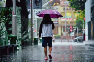 Rear‑view street‑level shot of a person walking along a rain‑soaked city sidewalk under an umbrella, capturing urban lifestyle, rainy weather atmosphere, and everyday city scenes.