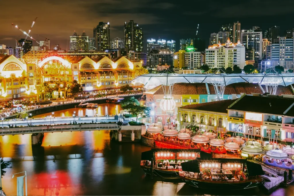 Wide-angle nighttime view of Clarke Quay and Boat Quay along the Singapore River, captured from an elevated vantage point, showing illuminated riverside buildings, bridges, reflections on the water, and vibrant nightlife activity.
