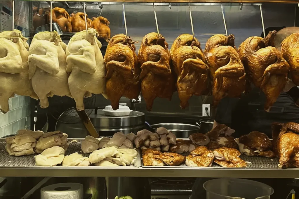 Wide-angle eye-level shot of a Singapore Hainanese chicken rice hawker stall display featuring rows of whole poached and roasted chickens hanging on metal hooks, sliced chicken pieces arranged on a stainless-steel counter below, and a busy open kitchen setup with cooking equipment in a traditional hawker centre environment.