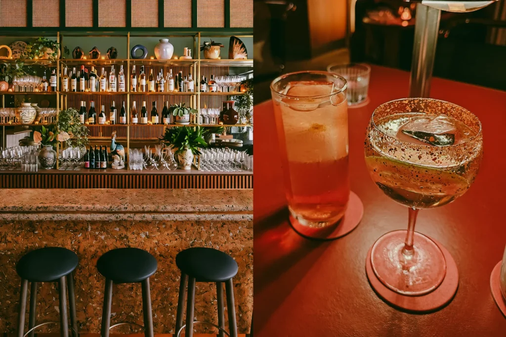 Left: An eye-level, straight-on wide shot of a stylish bar featuring a textured earthy terrazzo counter fronted by three minimalist black stools. The back bar is neatly arranged with brass shelves displaying rows of wine and liquor bottles, glassware, ceramic vases, and green indoor plants against a paneled wall. Right: A close-up, high-angle shot focusing on two iced drinks served on a smooth red table surface. In the foreground, a stemmed glass cocktail features a rim coated with dark spices and a lime wheel slice inside, while a tall highball glass filled with an amber-colored layered beverage and large ice cubes sits just behind it under warm, dim illumination.