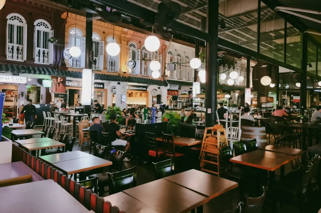 Wide‑angle interior shot of Cuppage Terrace in Singapore, featuring rows of wooden tables, hanging globe lights, multiple Asian restaurants, and diners seated under a covered arcade in the evening.