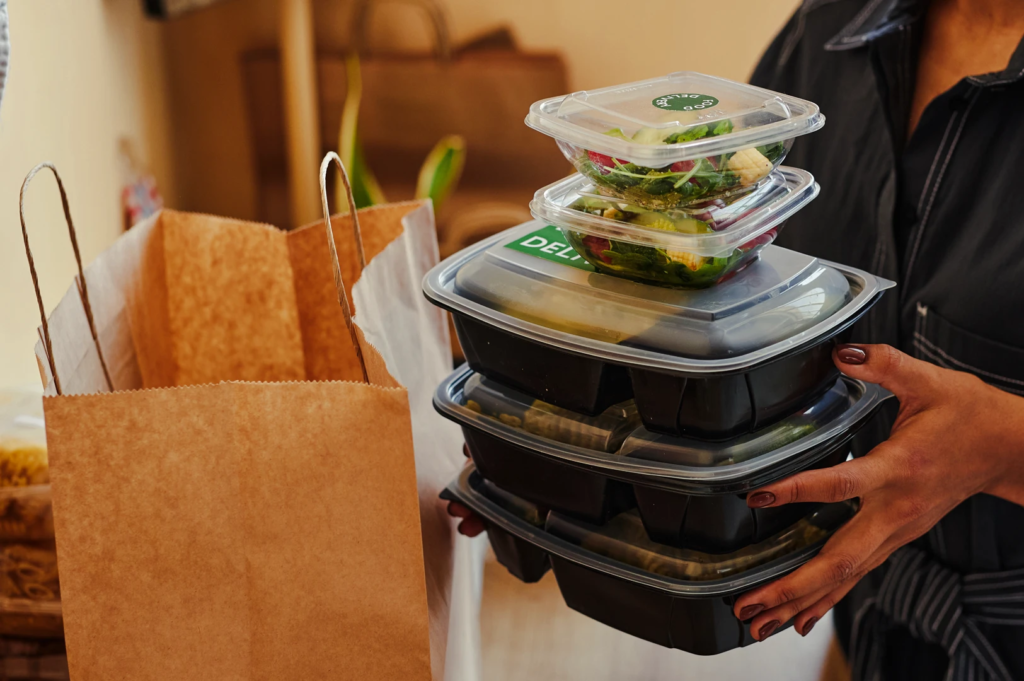 Stack of reusable meal prep containers filled with salads and cooked dishes being packed into a paper bag for takeaway.