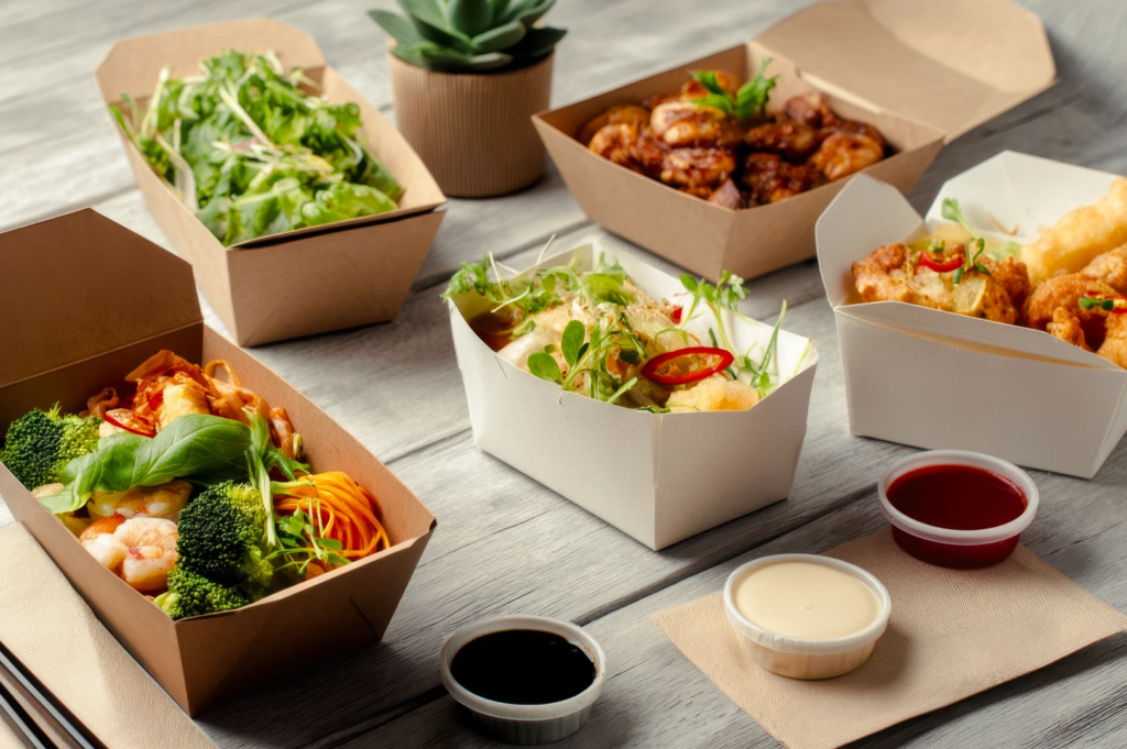 Assorted takeaway dishes in eco‑friendly cardboard containers, including noodles, salads, prawns, and fried items with dipping sauces on a wooden table.