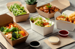 Assorted takeaway dishes in eco‑friendly cardboard containers, including noodles, salads, prawns, and fried items with dipping sauces on a wooden table.