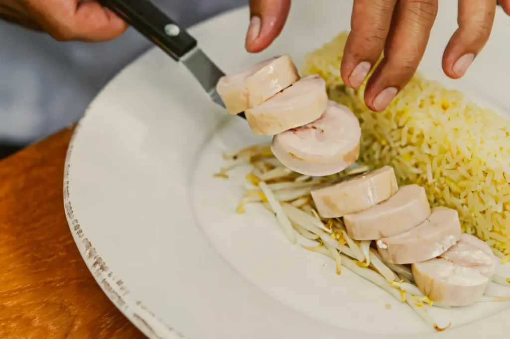 Top-down close-up shot of freshly sliced poached chicken being plated beside yellow chicken rice and bean sprouts, highlighting preparation of traditional Hainanese chicken rice.