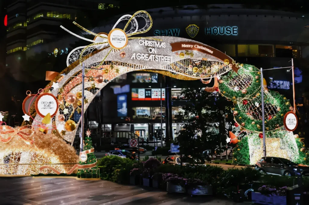Wide-angle night shot of Orchard Road in Singapore during the Christmas light-up, featuring illuminated festive arches, holiday ornaments, traffic, and surrounding buildings.