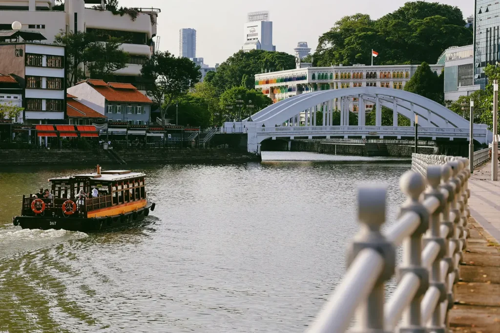 Mid-range daytime shot of Elgin Bridge spanning the Singapore River, photographed from a riverside railing perspective, with a bumboat on the water and historic buildings lining Boat Quay.
