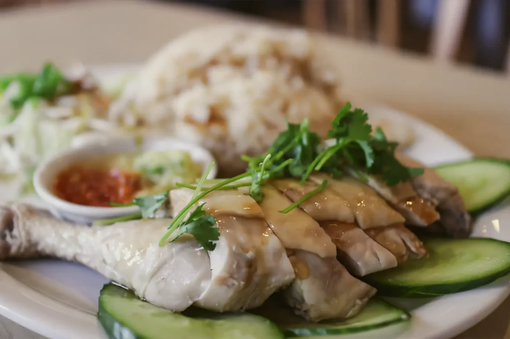Close-up slightly angled shot of sliced poached Hainanese chicken garnished with cilantro and served with cucumber, chili sauce, and fragrant rice on a white plate.