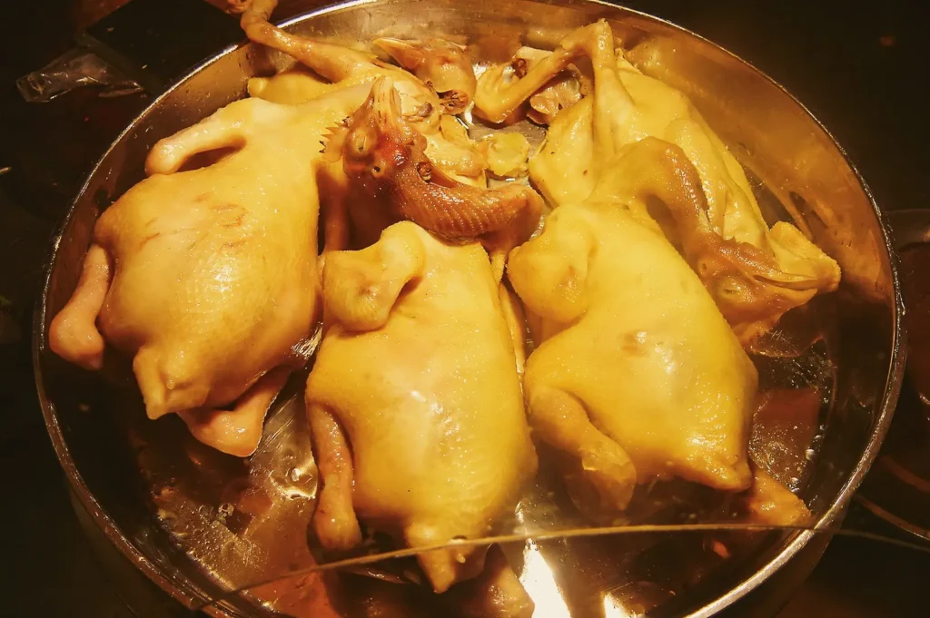 Top-down close-up shot of whole chickens simmering in a large metal pot of broth, illustrating the traditional poaching process for Wenchang chicken.