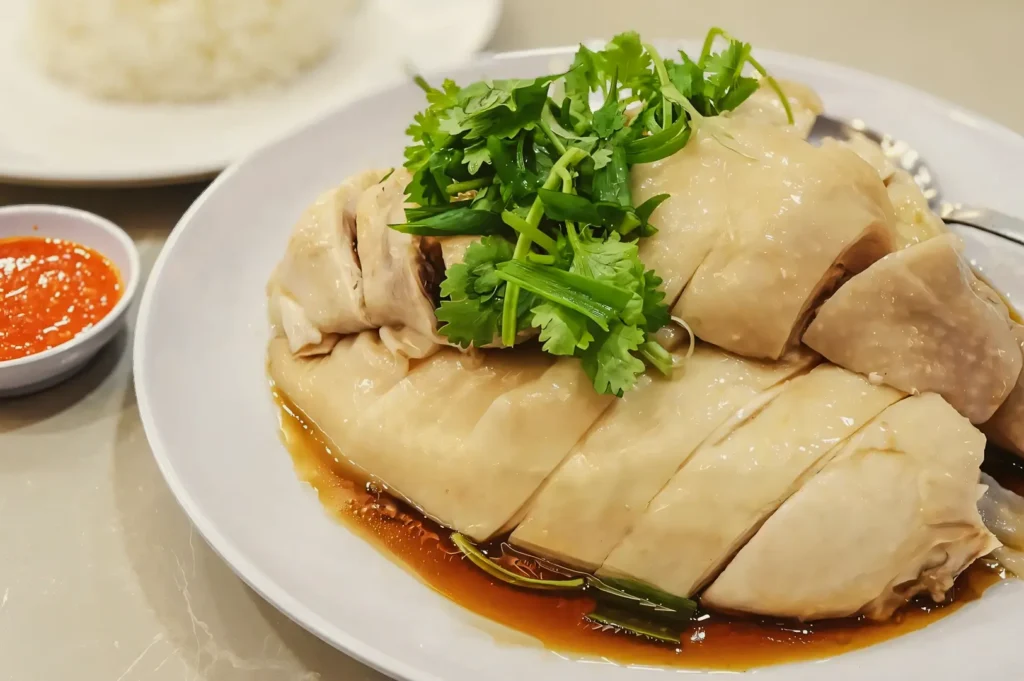 Close-up eye-level shot of classic Singapore Hainanese chicken rice with tender poached chicken glazed in soy sauce, garnished with fresh cilantro and served alongside chili sauce on a white plate.