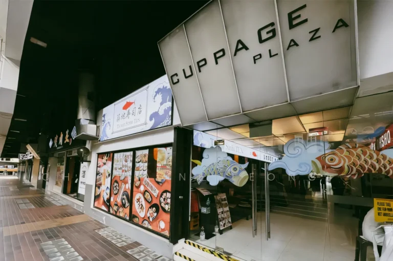 Low eye‑level exterior shot of Cuppage Plaza entrance along a pedestrian walkway, showing illuminated restaurant signage, Japanese food displays, glass doors, and a covered arcade in Singapore.