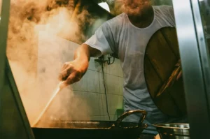 Close-up, slightly low-angle shot of a hawker cooking over a flaming wok, with steam and motion capturing the intensity of Singapore street food preparation.