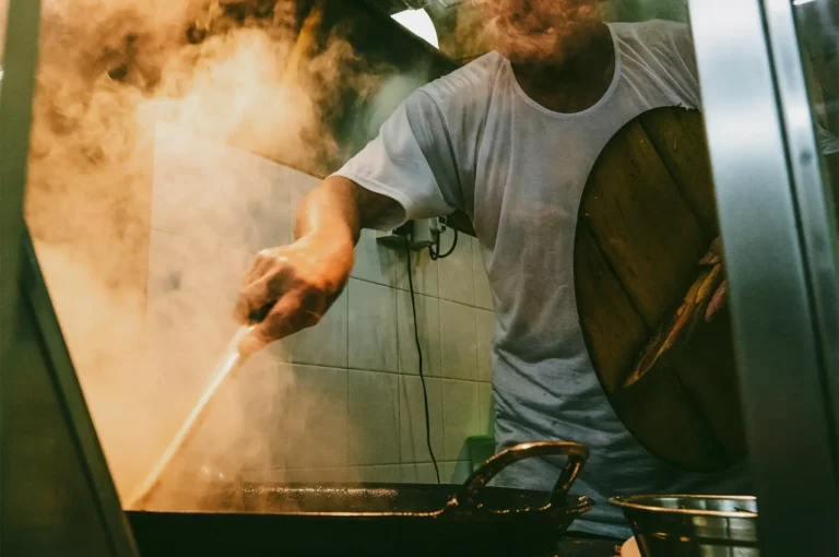 Close-up, slightly low-angle shot of a hawker cooking over a flaming wok, with steam and motion capturing the intensity of Singapore street food preparation.