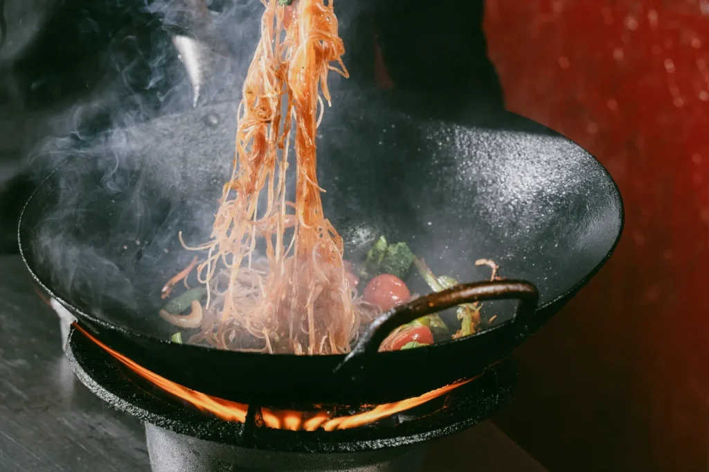 Close-up wok stir-fry shot with noodles, broccoli, cherry tomatoes, and peppers tossed over open flame, steam rising with wok hei aroma.