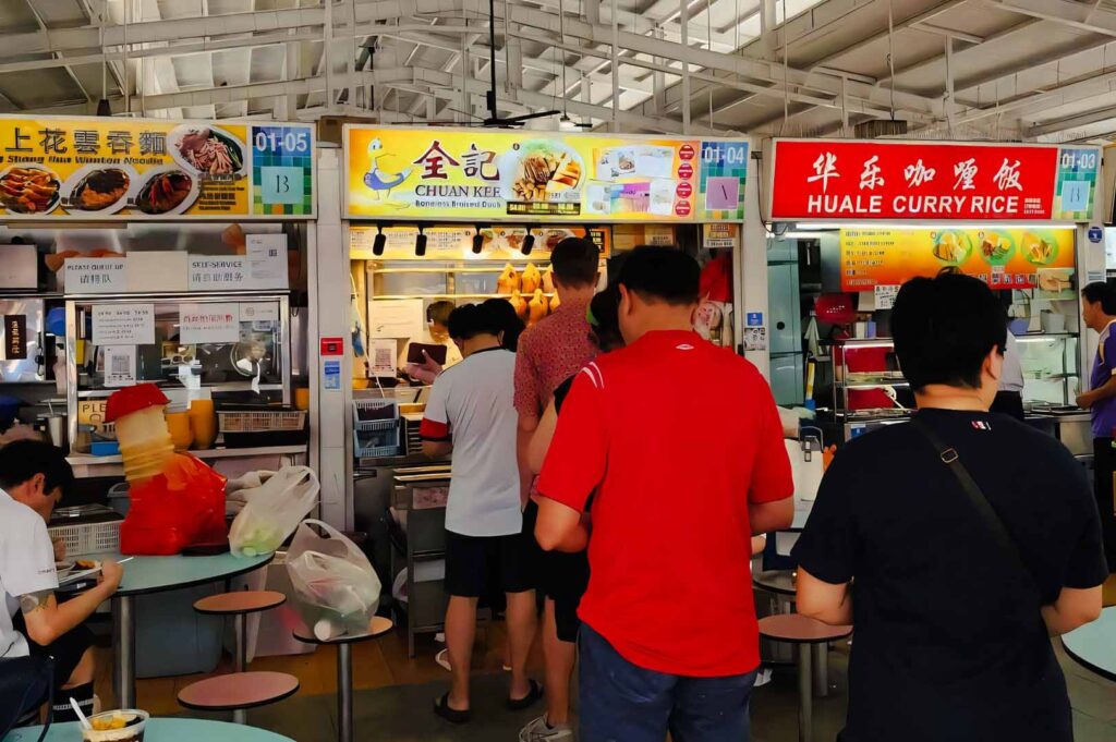 Queue of customers at a Singapore hawker centre ordering food from various stalls, including noodle and curry rice vendors