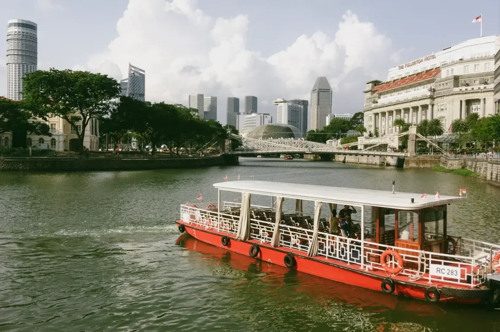 Wide-angle daytime view of a traditional bumboat cruising along the Singapore River, captured from a slightly elevated riverbank angle, with modern city buildings and riverside greenery in the background.