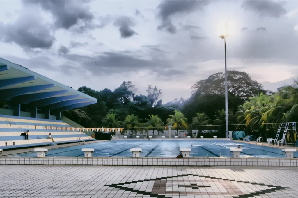Wide-angle, eye-level shot of an outdoor swimming pool in Bukit Timah, Singapore, with empty lanes, tiled deck, spectator stands, palm trees, and dramatic cloudy evening sky.