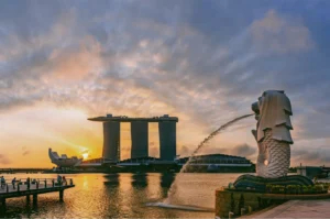 Wide-angle sunrise photograph of the Merlion statue spouting water toward Marina Bay, captured from a low waterfront angle with Marina Bay Sands silhouetted against a dramatic evening sky.