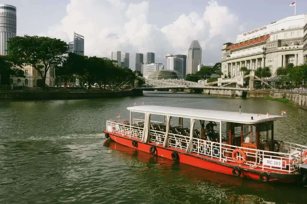 Wide-angle, eye-level shot of a red bumboat cruising along the Singapore River, with the city skyline, historic buildings, and waterfront greenery in the background.