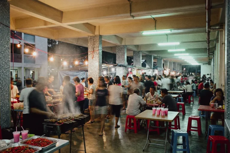 Wide‑angle eye‑level shot of a bustling void deck market set beneath a residential building, featuring food stalls, grilling stations, shared tables, and shoppers moving through an open concrete space, highlighting Singapore community markets and pop‑up street food culture.