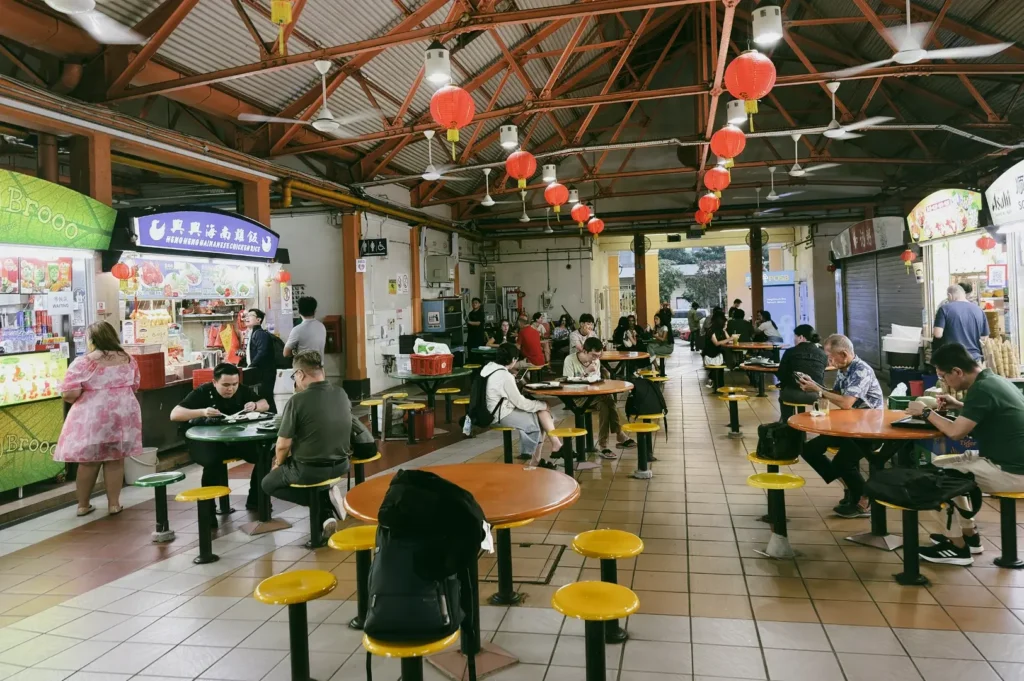 Wide-angle, eye-level interior shot of a Singapore hawker centre with communal tables, yellow stools, red lanterns, and diners enjoying neighborhood food.