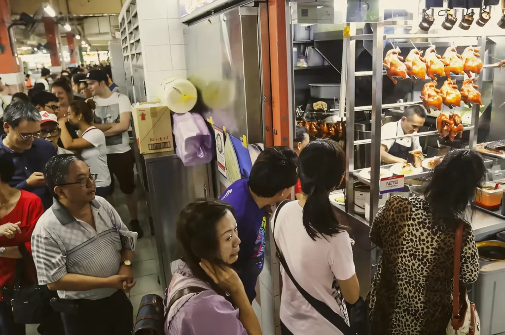 Wide-angle slightly elevated shot of a crowded Singapore hawker centre chicken rice stall, showing customers queueing beside an open kitchen with hanging roasted chickens and stainless-steel workstations.