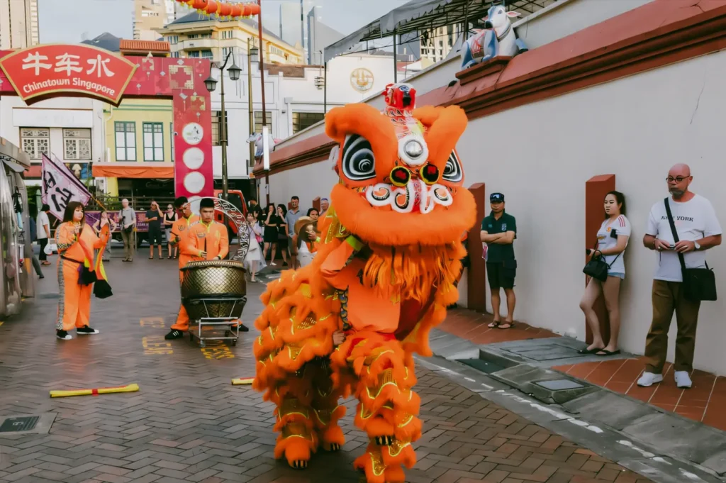 Eye-level wide shot of a vibrant lion dance performance during Chinese New Year in Chinatown, Singapore, with performers in an orange lion costume moving through a lantern-lined street as onlookers watch.