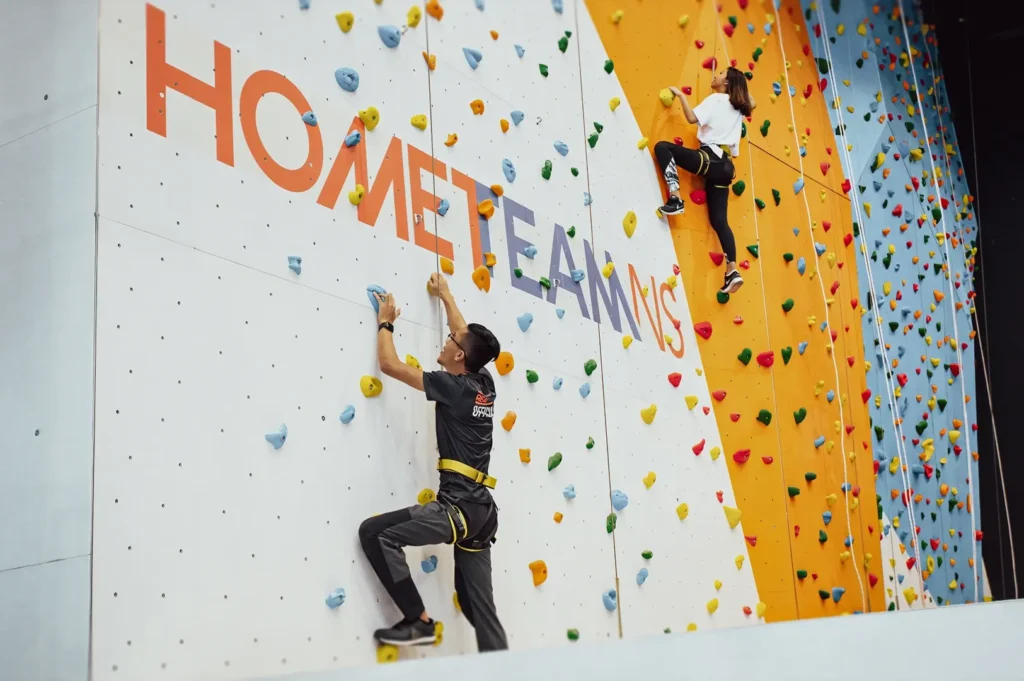 Wide‑angle action shot of climbers ascending a brightly colored indoor rock‑climbing wall with safety harnesses, showcasing fitness activities, climbing gyms, and active indoor recreation.