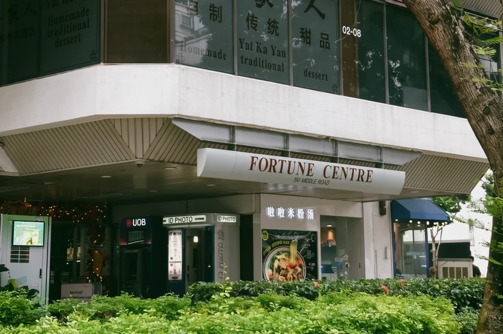“Wide-angle, eye-level exterior view of Fortune Centre at 190 Middle Road, showing the building entrance with a prominent ‘FORTUNE CENTRE’ canopy sign above a covered walkway. In the foreground, a landscaped hedge with dense green foliage runs along the front of the property."