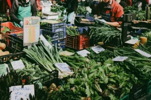 Eye-level wide-angle view of a busy farmers market stall filled with fresh seasonal vegetables, local vendors arranging leafy greens and organic produce, showcasing community market life and sustainable shopping