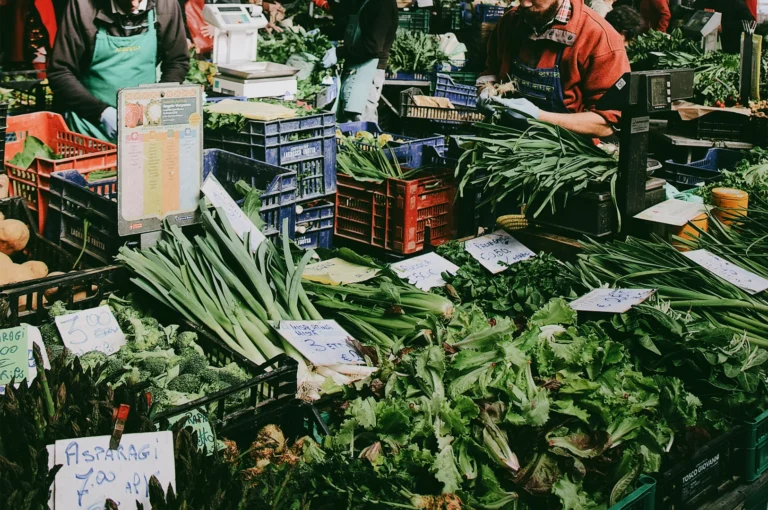 Eye-level wide-angle view of a busy farmers market stall filled with fresh seasonal vegetables, local vendors arranging leafy greens and organic produce, showcasing community market life and sustainable shopping