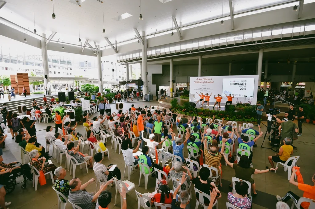 High-angle wide shot of a Singapore neighborhood community event at an HDB void deck plaza featuring residents participating in group activities and stage performances promoting social bonding.