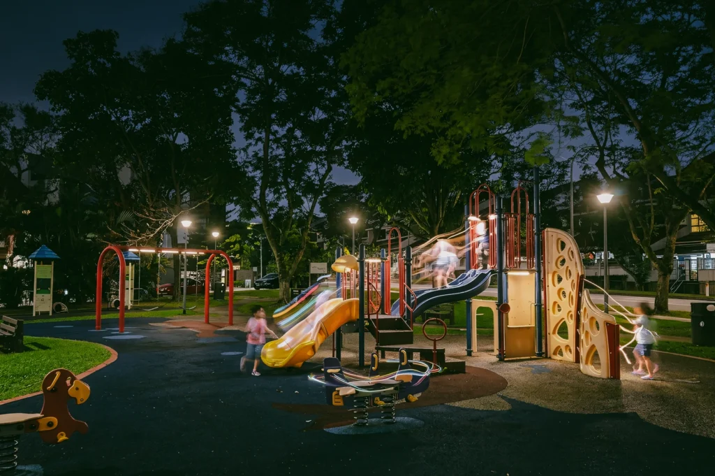 Wide-angle eye-level night shot of children playing on colorful slides in a neighbourhood playground, captured with motion blur under park lights in an urban residential area.