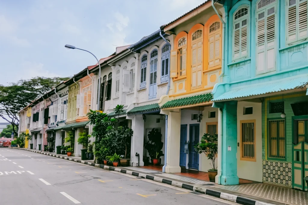 Street-level view of colorful heritage shophouses in a row featuring pastel facades, shuttered windows, and covered walkways along a quiet urban street, showcasing classic Southeast Asian architecture.