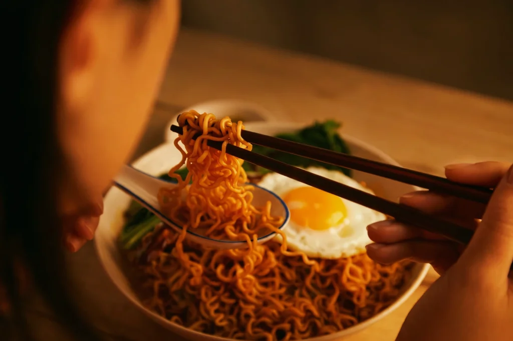 Close-up over-the-shoulder shot of chopsticks lifting Maggi mee noodles with fried egg in a bowl, warm lighting highlighting a cozy late-night Singapore supper ritual