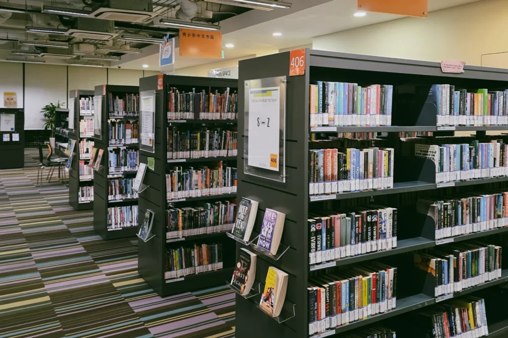 Eye-level wide-angle shot of modern Singapore community library bookshelves (section 406) filled with fiction and non-fiction, showcasing organized aisle layout and inclusive public reading space