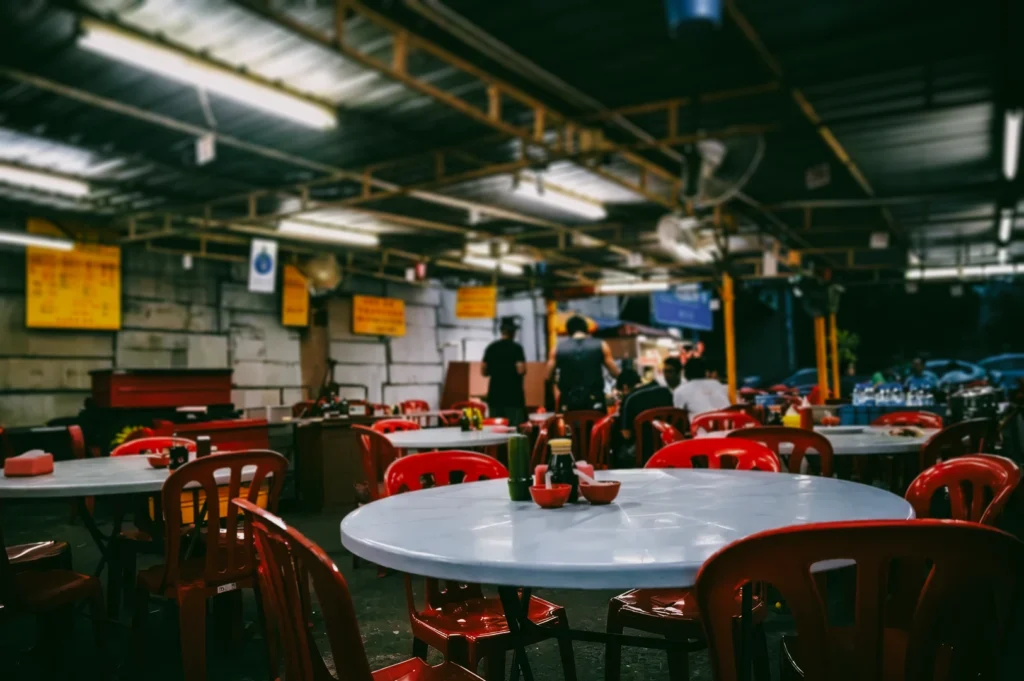 Wide-angle, eye-level shot of an open-air Singapore hawker centre with red plastic chairs and round tables under fluorescent lighting, showcasing casual street food dining culture, shared seating, and nostalgic local kopi shop atmosphere.