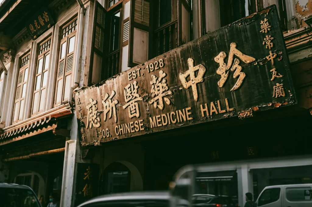 Weathered heritage Chinese medicine hall signboard with vintage typography reading “Chinese Medicine Hall Est. 1928” on a traditional shophouse facade, captured in a slightly low-angle street shot emphasizing nostalgic architecture and old Singapore cultural landmarks.