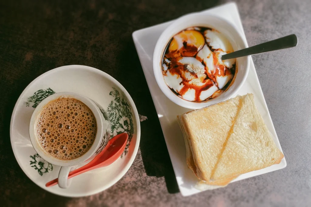 Overhead flat lay of a traditional kopi and kaya toast breakfast featuring a cup of frothy local coffee (kopi) in a ceramic bowl with a red spoon, soft-boiled eggs drizzled with soy sauce and pepper, and stacked kaya toast on a white plate.