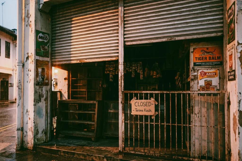 Vintage Southeast Asian corner shop with metal shutters partially closed, weathered walls, and a “Closed – Terima Kasih” sign alongside retro Milo and Tiger Balm advertisements, captured in a cinematic eye-level street angle highlighting nostalgia and disappearing local businesses.