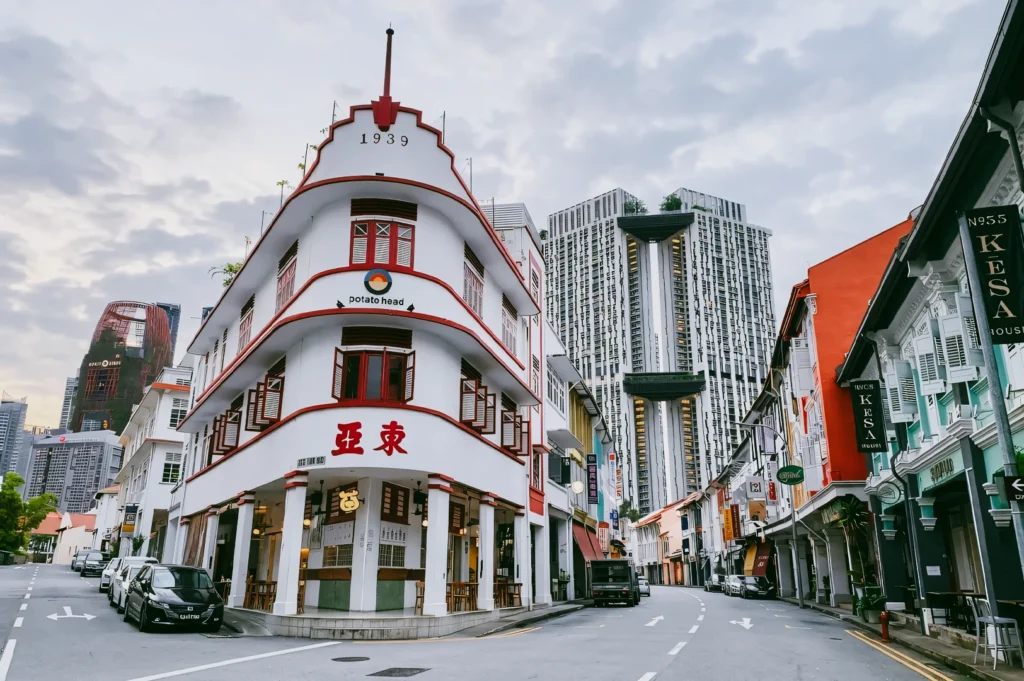 Low-angle wide shot of curved heritage shophouse on Keong Saik Road, Singapore, with red-trim facade, early morning light, and modern skyscrapers in background highlighting old vs new cityscape