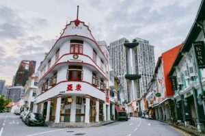 Low-angle wide shot of curved heritage shophouse on Keong Saik Road, Singapore, with red-trim facade, early morning light, and modern skyscrapers in background highlighting old vs new cityscape